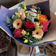Load image into Gallery viewer, A vibrant luxury flower posy wrapped in dark purple and brown paper, featuring a red rose, orange and yellow gerbera daisies, pink astilbe, and green foliage, displayed on a small wooden stool. delivered by Bits and Buds local florist in leighton Buzzard