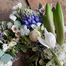 Load image into Gallery viewer, Floral arrangement with white flowers, green leaves, and a speckled egg on a wooden surface