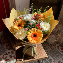 Load image into Gallery viewer, A vibrant not artificial fresh flower bouquet wrapped in gold paper, featuring orange and white gerbera daisies, white roses, and green foliage, displayed on a small wooden stool in a home setting.
