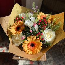 Load image into Gallery viewer, A vibrant autumn mixed flower bouquet wrapped in brown kraft paper, featuring orange and yellow gerbera daisies, white roses, small pink blossoms, and green foliage, displayed on a small wooden stand.
