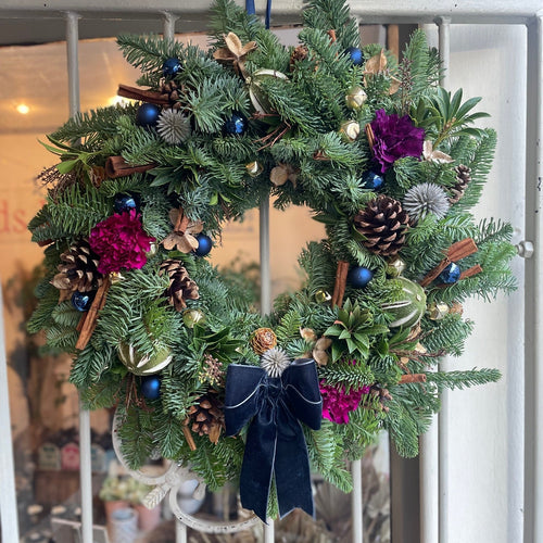 Decorative Christmas wreath with pine cones, flowers, and a blue bow on a white door.