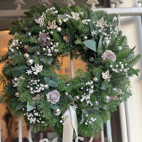 natural white christmas door weath with silver bells fresh babys breath flowers wooden doves and white ribbons on a luxurious spruce greenery base from bits and buds florist christmas shop in leighton buzzard 