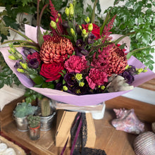 Load image into Gallery viewer, Bouquet of blood red and black flowers with a black lace ribbon and purple wrap on a wooden table with plants in the background
